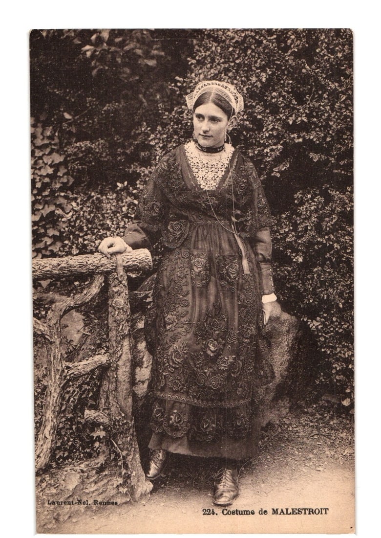 Sepia postcard depicting a woman in traditional Malestroit costume, by Laurent-Nel, Rennes.: A sepia-toned photographic postcard depicts a young woman in elaborate traditional costume, standing outdoors against dense foliage. She looks towards the viewer's right. Her attire includes a dark la