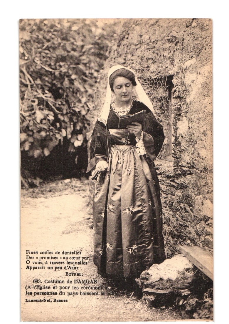 Woman in Damgan Traditional Costume with Book, French Cultural Postcard, Early 20th Century: A young woman in a traditional costume stands outdoors, looking down at an open book held in her hands. She wears a dark dress with a long, full skirt and a white lace coif covering her head, with a v