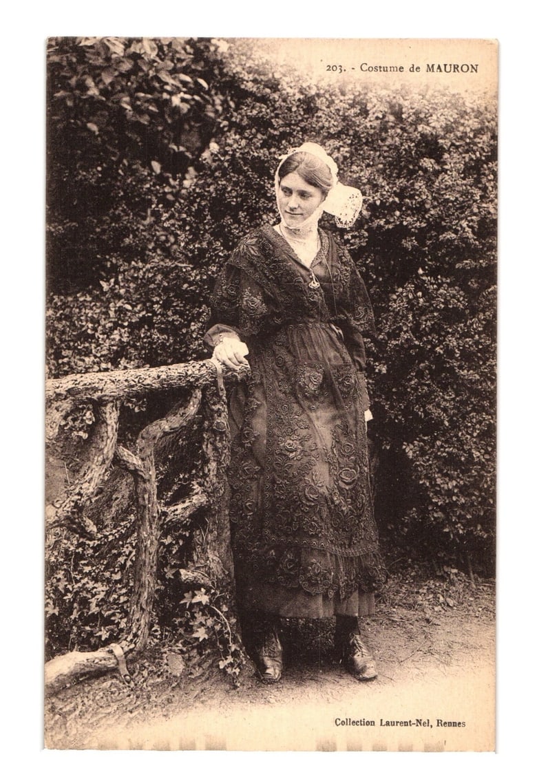 Young Woman in Traditional Mauron Costume, Rennes, France, Sepia Photographic Postcard: A young woman, depicted full-length, stands outdoors, leaning against a rustic, gnarled railing. She wears a dark, intricately patterned traditional dress, possibly lace or embroidered, featuring long