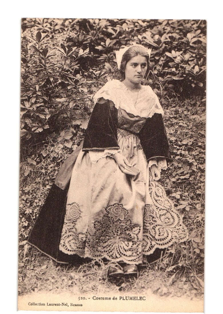 Early 20th Century Postcard: Woman in Plumelec Traditional Costume with Lace and Velvet, France: A young woman is seated amidst dense foliage, looking slightly to her right. She wears an elaborate traditional costume, which includes a white head covering, a white blouse with lace-trimmed sleeves,