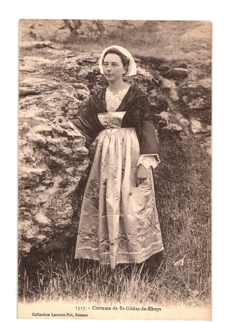 Full-length photographic postcard of a woman in traditional French costume, St-Gildas-de-Rhuys: A full-length photographic portrait depicts a woman in traditional French costume. She wears a white cap or headdress, a dark long-sleeved garment with lace trim over a lighter blouse, and a long, lig