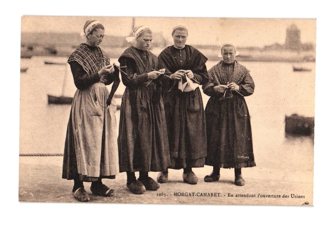Four Breton Women Knitting on a Pier in Traditional Attire, Morgat-Camaret postcard: Four women, dressed in traditional attire comprising long dark dresses, white aprons, patterned shawls, and white head coverings, stand on a pier or waterfront. Three of the women are actively engaged