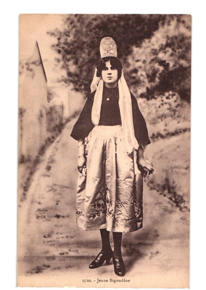 Young Bigouden Woman in Traditional Costume, Studio Portrait Real Photo Postcard, Early 20th Century: A full-length, sepia-toned photographic portrait captures a young woman in traditional attire, standing centrally against a dark, possibly painted studio backdrop depicting foliage and a path. She wea