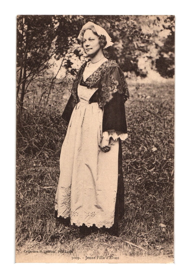 Early 20th Century sepia postcard of a young girl in traditional Elven costume.: A young woman stands outdoors, dressed in traditional attire, with trees and brush in the background. She wears a white bonnet, a dark lacy shawl or cape over a blouse, and a long, light-colored apron