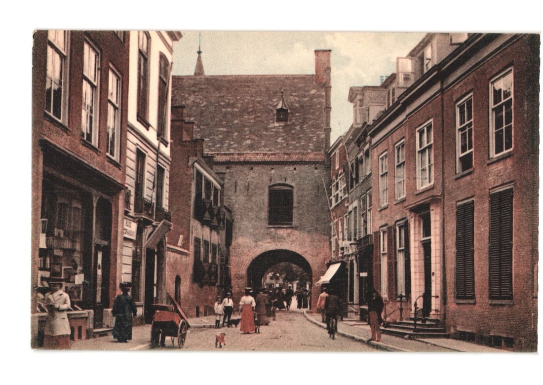 Early 20th Century Street View of The Hague's historic Gevangenpoort, Netherlands postcard: A street scene depicts a prominent, multi-story brick or stone archway structure, the Prison Gate, at the end of a cobblestone street. Buildings with multiple windows and shutters line both sides of t