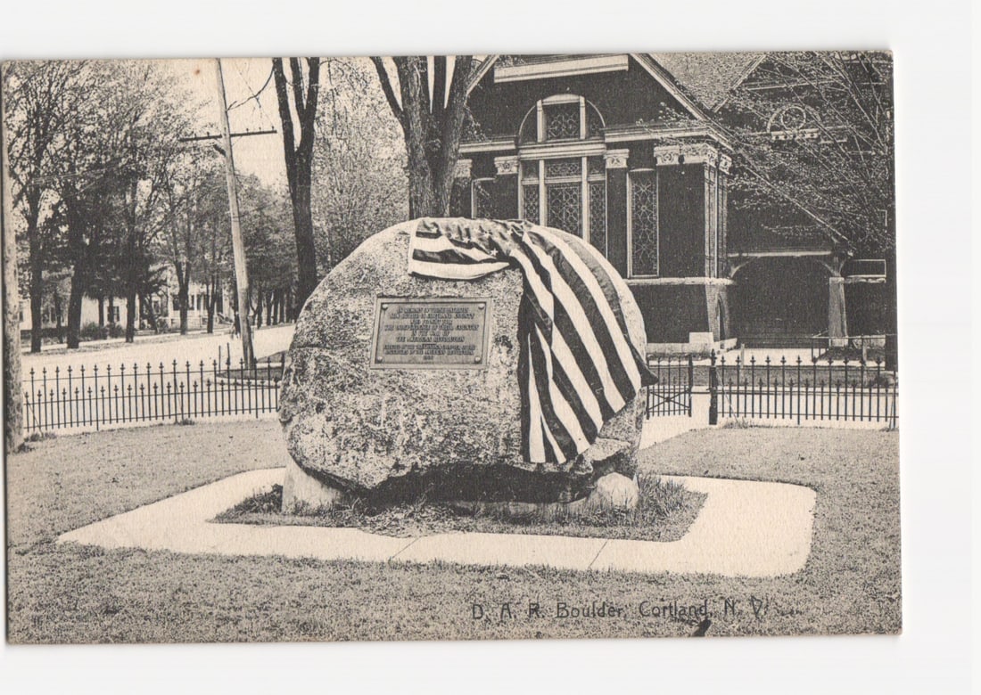 D.A.R. Boulder Monument, Cortland, NY, Early 20th Century Photographic postcard.: A large, irregular boulder, draped with an American flag, stands centrally on a manicured lawn. The boulder is positioned on a concrete base, enclosed by a low metal fence. A bronze plaque is affixed 