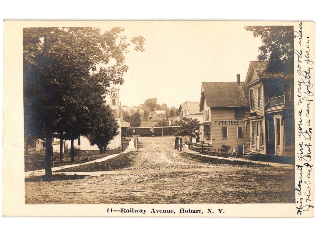 Railway Avenue, Hobart, N. Y. street view with furniture store, church, and train, 1909 postcard.: A sepia-toned real photo postcard (RPPC) captures a street scene on Railway Avenue in Hobart, New York. The unpaved street extends towards the background, bordered by lush trees on the left and severa