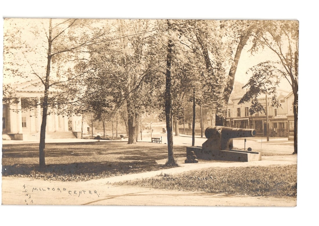 Milford, Connecticut, Town Green with Cannon Monument and Neoclassical Building, circa 1912: A sepia-toned Real Photo Postcard depicts a town green with a cannon monument in the foreground. The large, dark cannon rests on a stone base, positioned right of center. A curving path leads from the