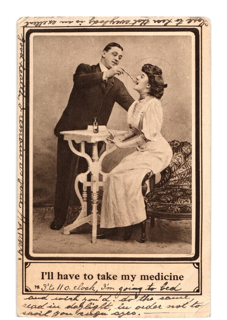 Early 20th-century sepia-tone postcard depicting a man feeding medicine to a seated woman.: A sepia-tone photographic image depicts a man in a dark suit standing and holding a spoon to the mouth of a woman seated in an ornate upholstered chair. The woman wears a light-colored dress and appea