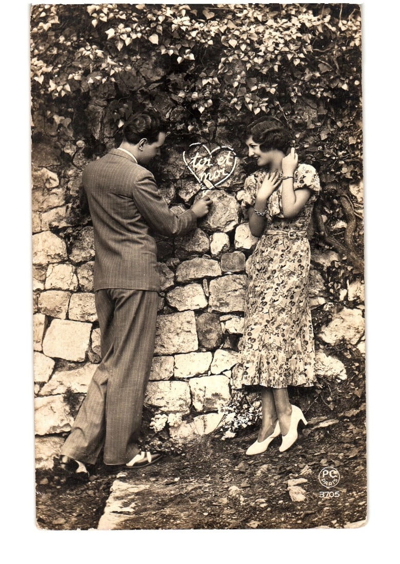 Early 20th-century French romantic postcard: a couple with a 'You and I' heart on a stone wall.: A man in a suit and a woman in a patterned dress stand beside a stone wall, partly covered by ivy. The man points to a heart-shaped object on the wall containing the text 'you and I'. The woman looks 