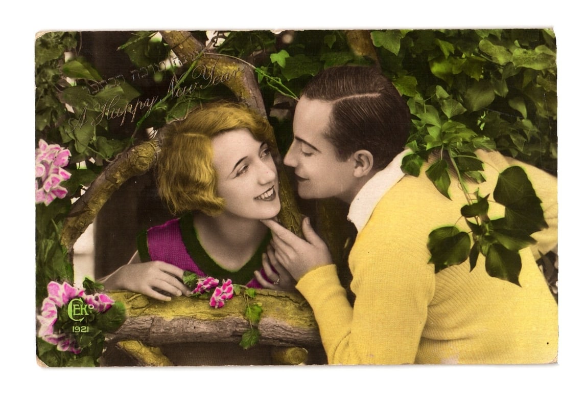 1921 Hand-Colored Photo Postcard of Young Couple in Foliage, with 'Happy New Year' Greeting: A hand-colored Real Photo Postcard depicts a young man and woman in an intimate pose, framed by green foliage and tree branches. The woman, with short blonde hair, wears a magenta top with green trim 