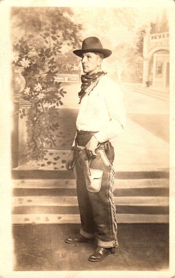 Sepia tone real photo postcard of a cowboy in studio Western attire, early 20th century.: A young man in cowboy attire is presented in a full-length sepia-tone studio portrait. He wears a dark cowboy hat, a patterned bandana, a light-colored long-sleeve shirt, dark chaps with fringe over t