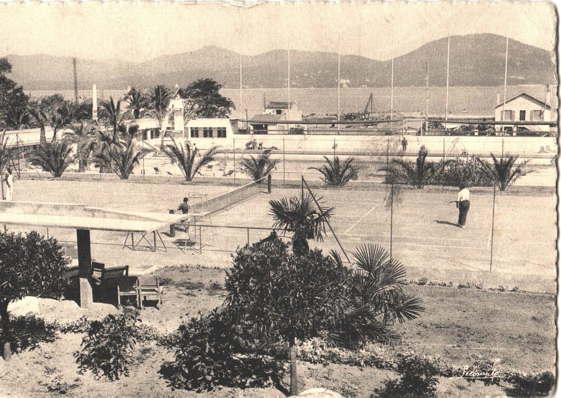 Mid-20th Century Saint-Tropez, France, postcard featuring tennis court and coastal vista: An elevated photographic view captures a tennis court with a coastal landscape in the background. Players are present on either end of the court, one seated near the net and another standing with a ra