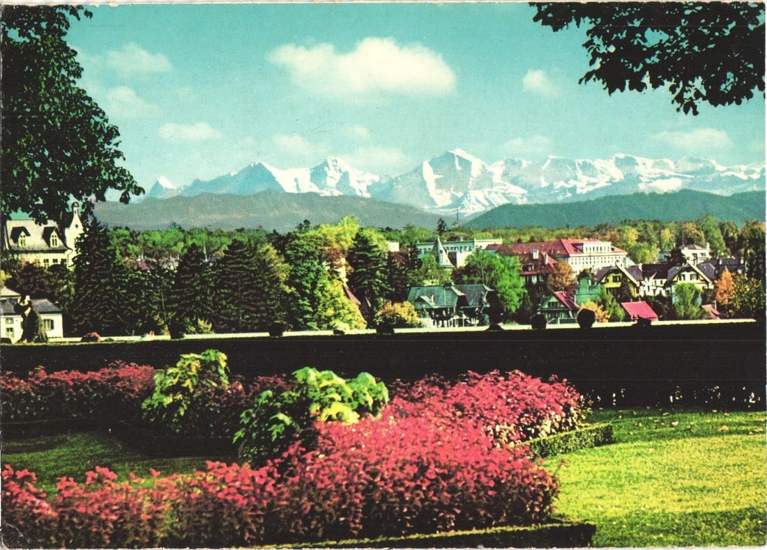 Scenic postcard depicting Bern, Switzerland, and the distant Alps from Bundesterrasse viewpoint.: A panoramic landscape view captures a broad vista from an elevated perspective. In the foreground, a well-maintained garden displays vibrant red and green foliage, separated from the middle ground by 