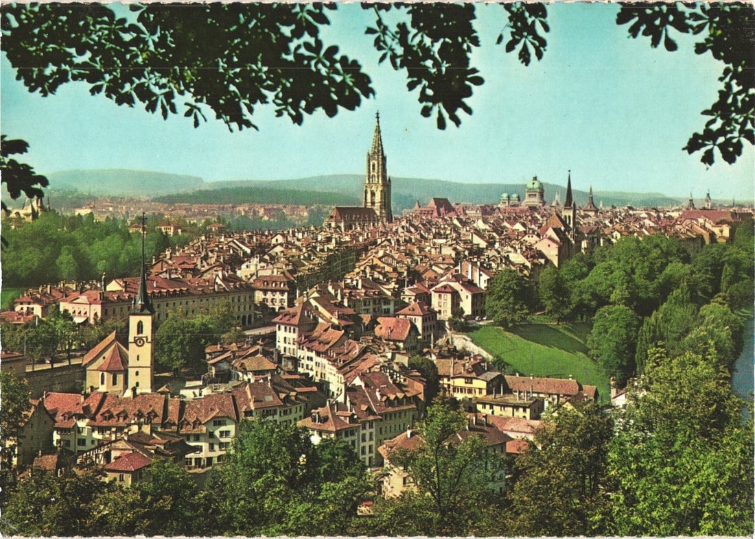 Historic Berne City Panorama, Switzerland, Featuring Cathedral Spire and Old Town Postcard: A panoramic cityscape featuring the Swiss capital of Berne, viewed from an elevated position. Dark green foliage partially frames the top edge of the image, looking down onto a densely built urban are