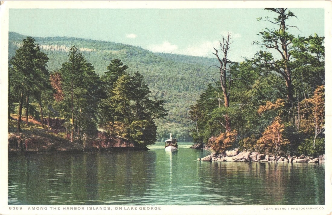 Boat Navigating Among Harbor Islands on Lake George, early 20th Century Phostint postcard: A small white motorboat with a cabin navigates a narrow passage of water, flanked by densely forested islands with rocky shores. Coniferous and deciduous trees cover the terrain, with larger hills vis