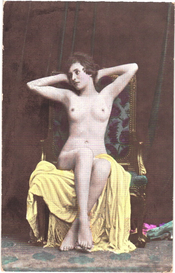 Early 20th-century hand-colored postcard of a nude woman seated in a studio setting.: A nude woman is depicted seated in an ornate, upholstered armchair, positioned against a plain, dark background. Her body is angled slightly to the right, with her head turned to face the right. Her a