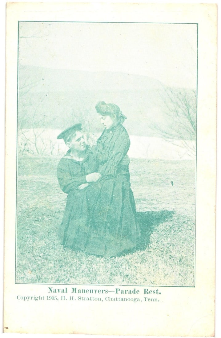 Monochrome postcard depicting a sailor and woman during "Parade Rest" in Chattanooga, 1905.: A monochrome image depicts a sailor in naval uniform kneeling, embracing a woman in a long dress, both looking at each other. They are positioned outdoors on a grassy field, with faint outlines of hil