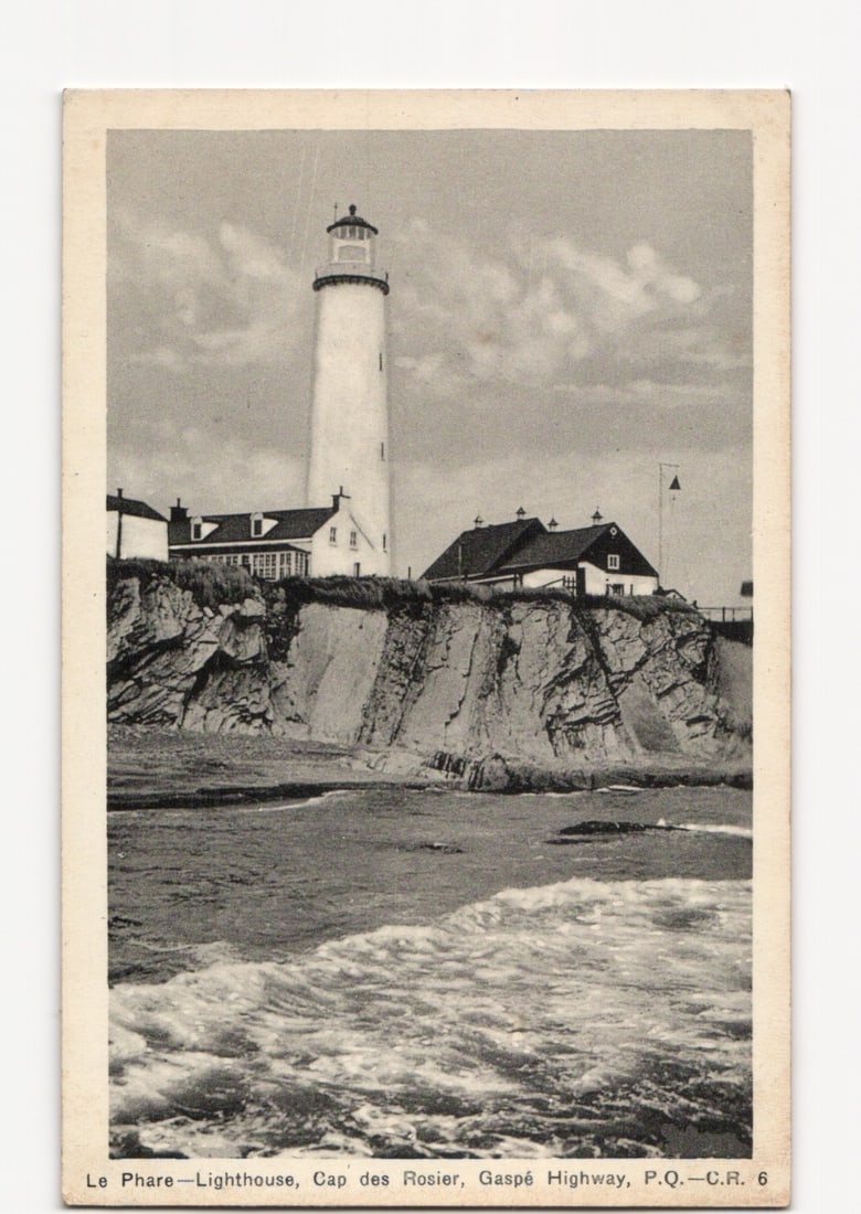 Le Phare Lighthouse, Cap des Rosier, Gaspe Highway, Quebec, Canada photographic postcard.: A tall, white, cylindrical lighthouse with a lantern room at its apex stands on a rocky cliff overlooking a body of water with breaking waves. Several ancillary buildings with pitched roofs are situat
