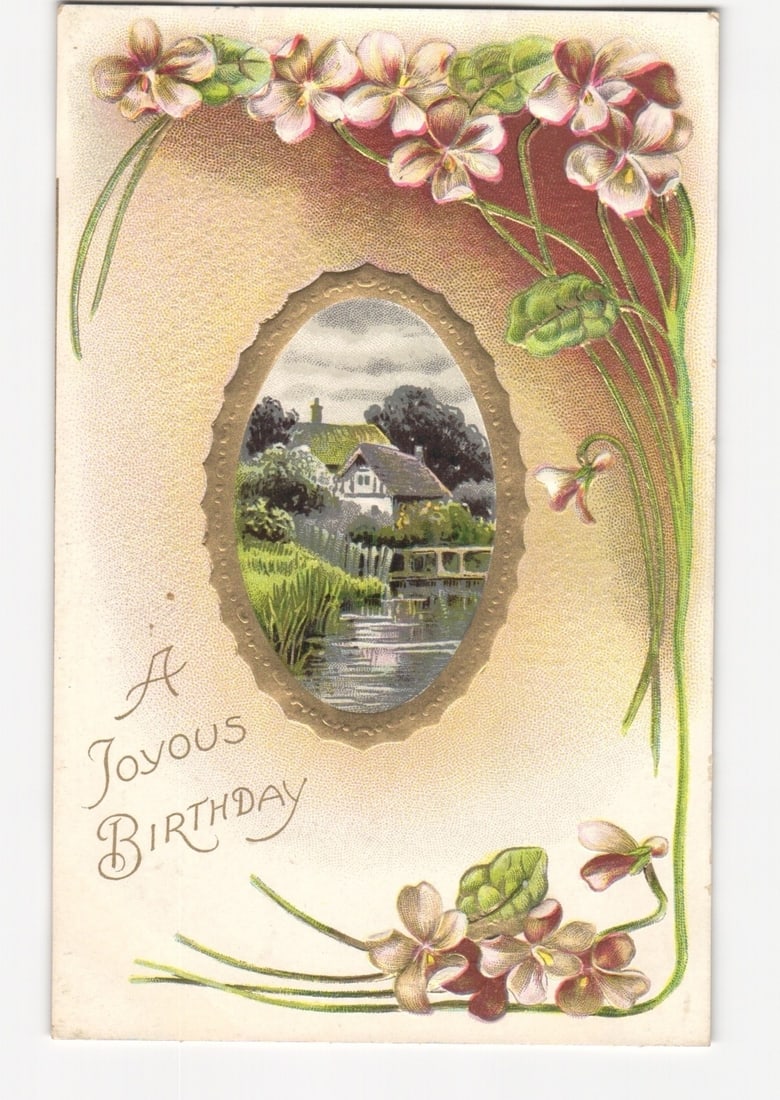 Illustrated Greeting Postcard: Cottage by Stream, Floral Violets, 'A Joyous BIRTHDAY', Printed in: A central oval vignette depicts a cottage situated by a flowing stream with a small bridge, surrounded by extensive trees and greenery, framed by a delicate gold border. The main image is flanked by d