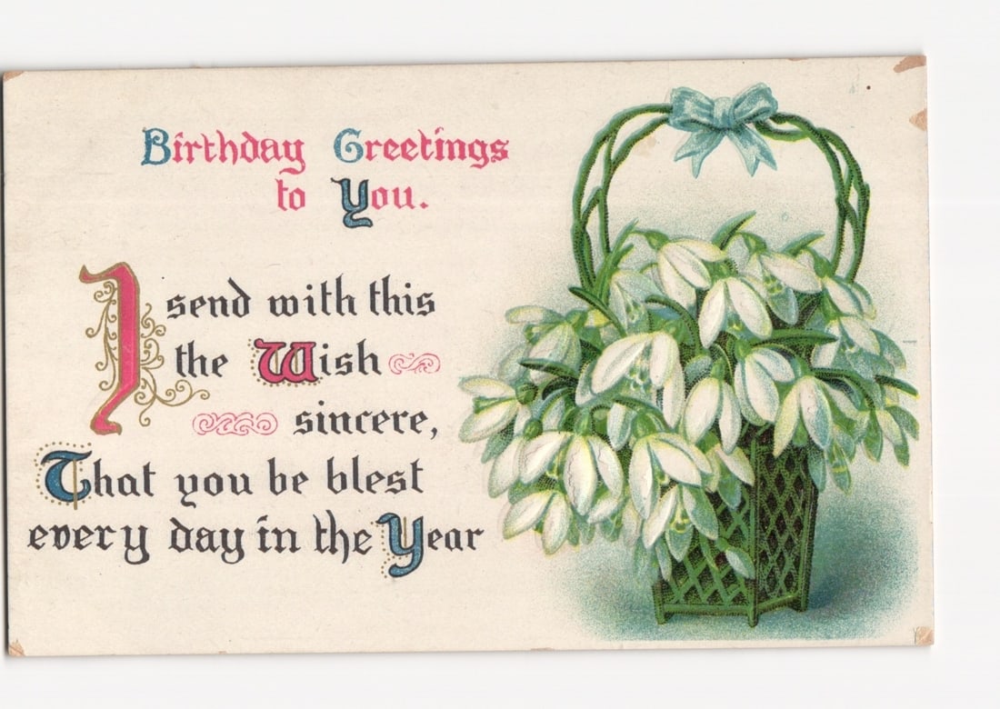 Early 1900s Birthday Greetings postcard with Snowdrop Flowers in a Decorative Basket: A chromolithograph postcard depicts a decorative woven basket overflowing with white snowdrop flowers, adorned with a light blue bow on its handle, positioned on the right side against a light backgro