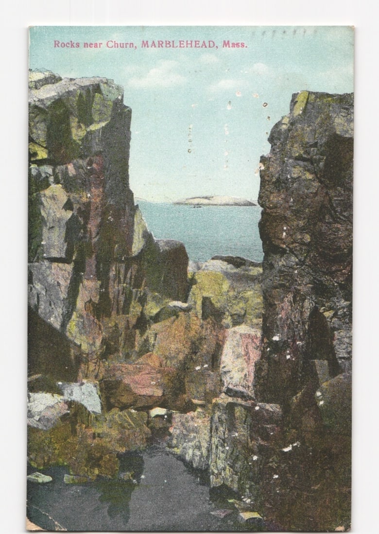 Colorized photographic postcard of rugged rocks near Churn overlooking the ocean, Marblehead, Mass.: A colorized photographic postcard captures a dramatic coastal scene, focusing on two towering, craggy rock formations that frame a view of the ocean. The rocks, mottled with green and brown hues, rise
