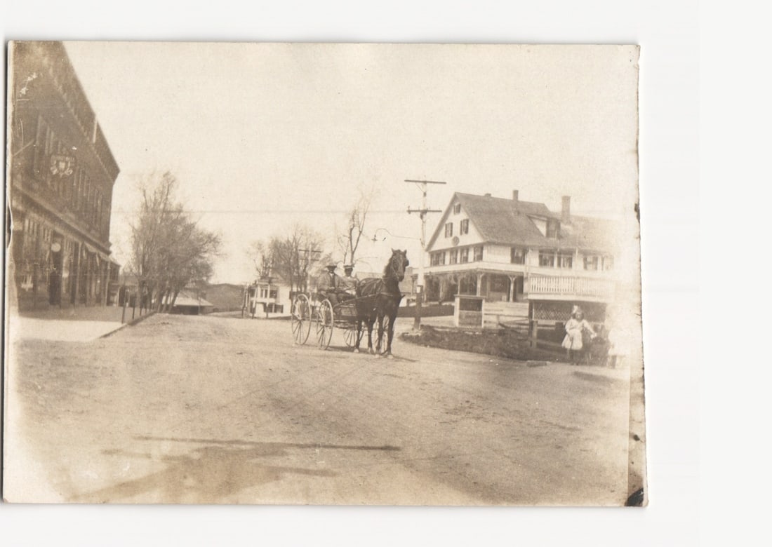 Village Street Scene with Horse-Drawn Carriage and Buildings, Early 1900s Real Photo postcard: A street scene features a horse-drawn carriage occupied by two individuals, positioned prominently in the center of an unpaved road. The horse faces right, harnessed to the carriage. To the left, a la
