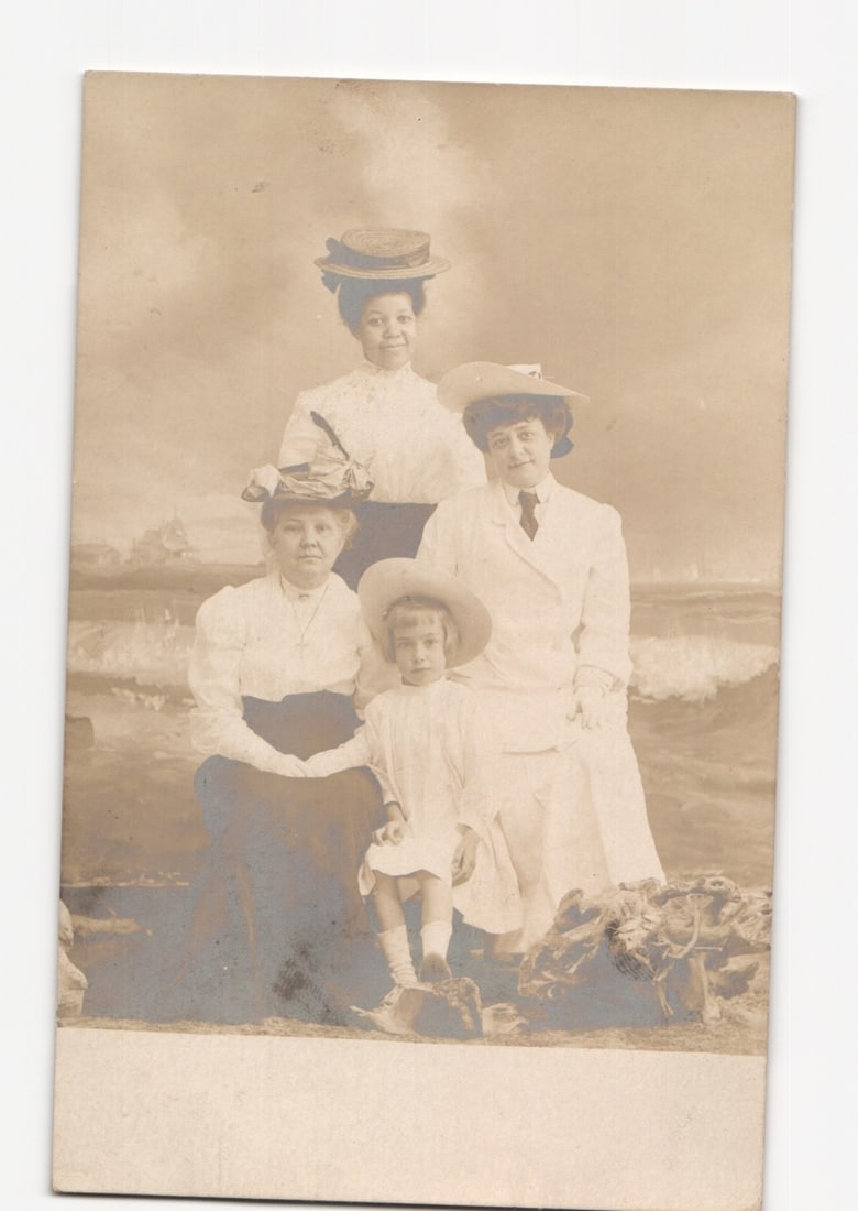 Studio postcard of three women and a child posed against a painted seaside backdrop, c. 1910.: A studio portrait depicts three adult women and one young child. The figures are arranged against a painted backdrop portraying a dynamic seaside scene with crashing waves, rocks, and a distant ship. 