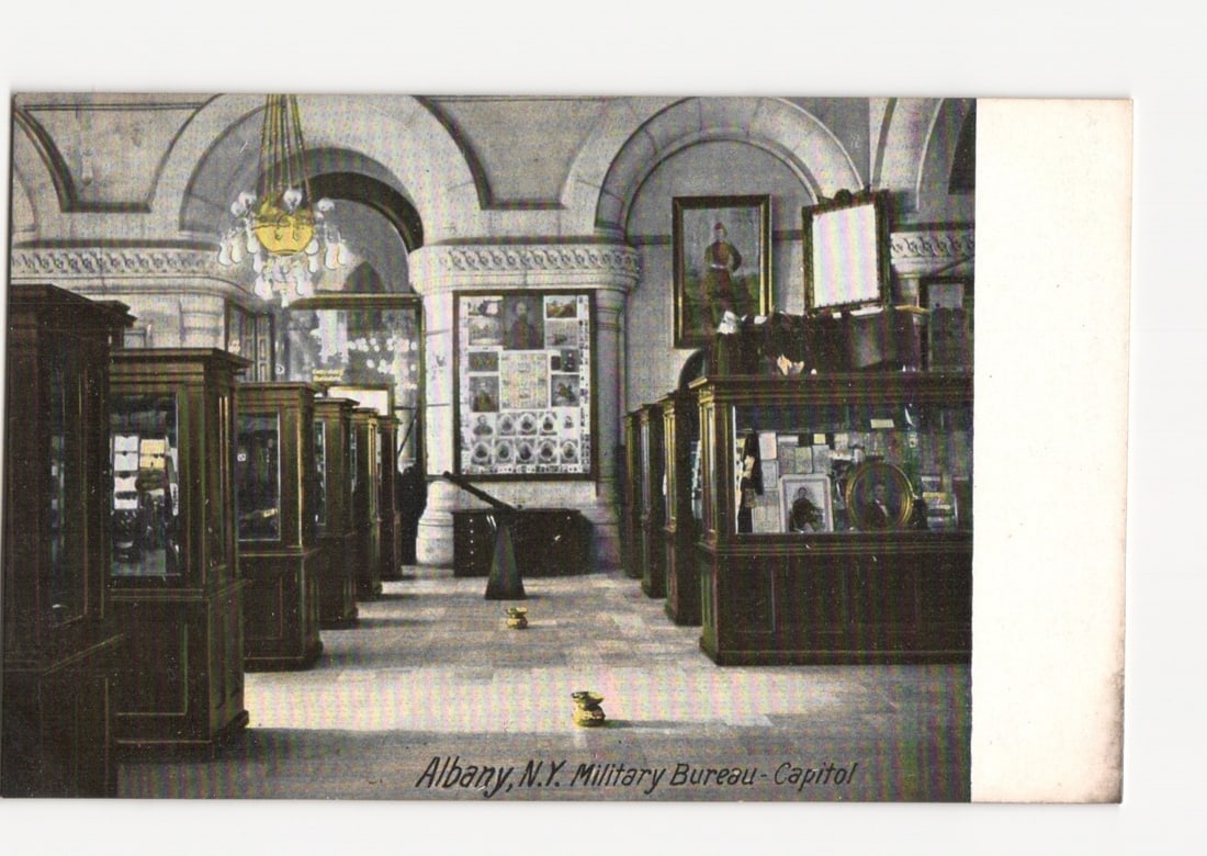 Albany, N.Y. Military Bureau - Capitol Interior with Display Cases, early 20th Century Postcard: An interior view depicts the Military Bureau within the Capitol building in Albany, New York. The scene shows a large hall with arched ceilings and pillars, illuminated by a chandelier. Numerous dark 