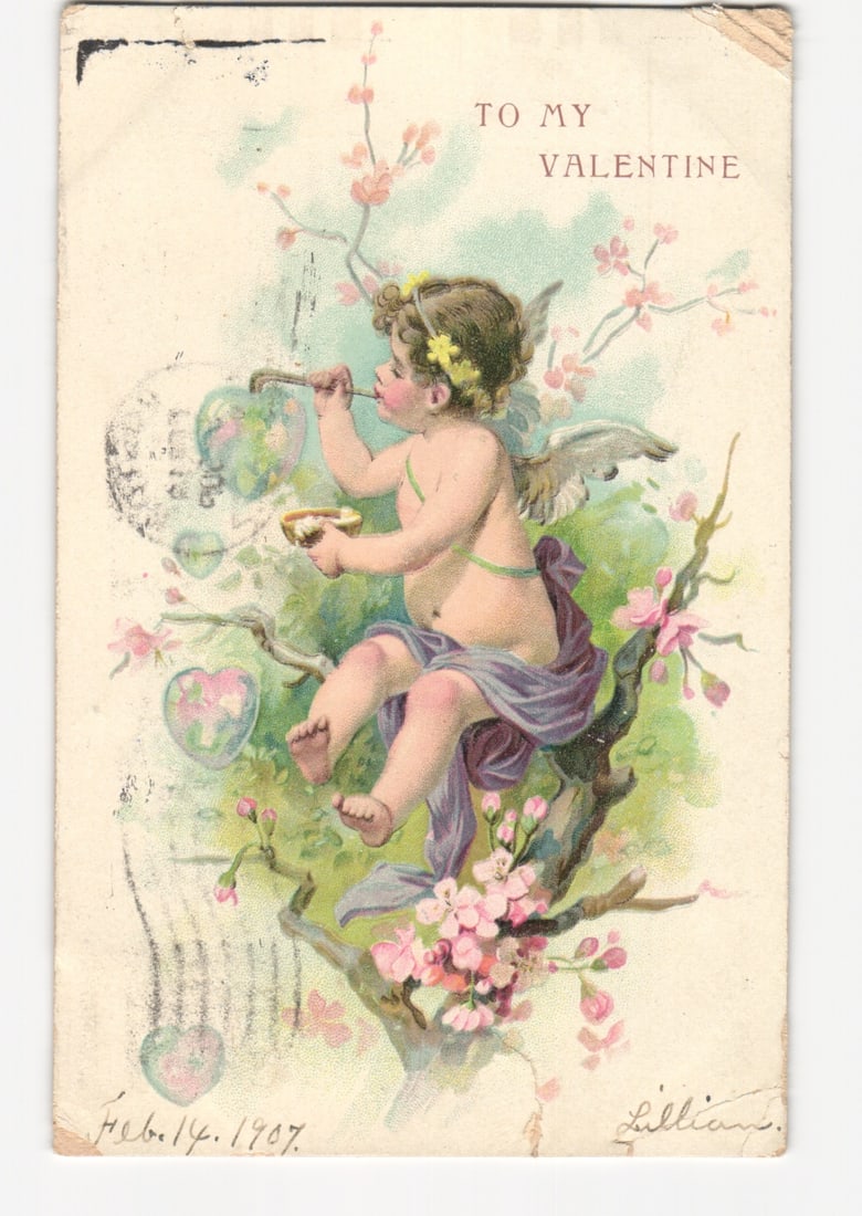 Vintage 1907 Valentine postcard featuring a winged cherub blowing heart bubbles in a flowering tree: A winged cherubic figure with blonde hair, wearing a yellow floral wreath, is seated in the branches of a flowering tree. The figure, partially draped in purple fabric and with a green ribbon around i