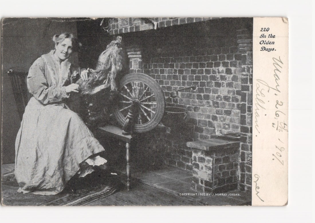 Vintage American Woman with Spinning Wheel by Fireplace Photographic Postcard, 1905-1907: A black and white photographic image depicts a woman seated beside a spinning wheel in an indoor setting. She wears a long, light-colored dress and smiles towards the viewer. A brick fireplace is visi