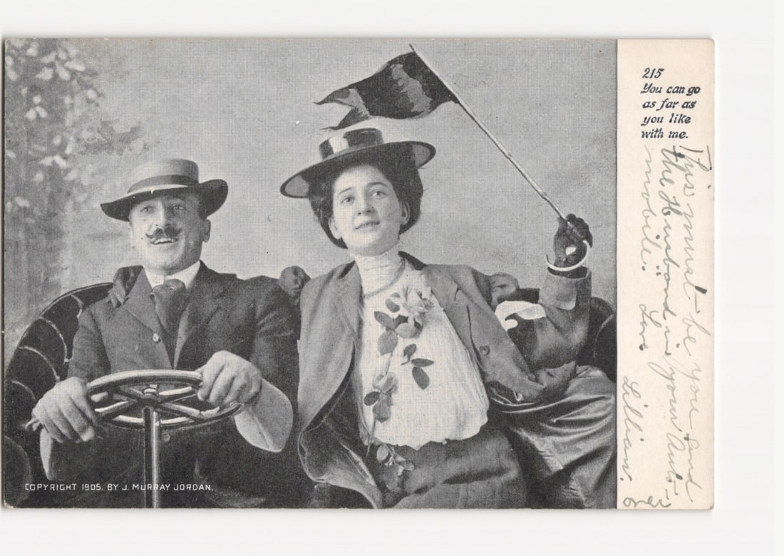 1905 Man and Woman in Early Automobile with Flag, Photographic Postcard by J. Murray Jordan: A man and a woman are seated in what appears to be an automobile. The man, on the left, has a mustache and wears a hat, suit, and tie, with his hands on a steering wheel. The woman, on the right, wear