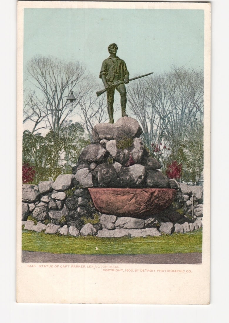 Statue of Captain Parker, Lexington, Massachusetts, an early 20th century photochrome postcard.: A colorized photograph depicts a statue of a man, identified as Captain Parker, standing on a rocky base. The figure wears period clothing and holds a rifle. The statue is centrally framed, with bare 