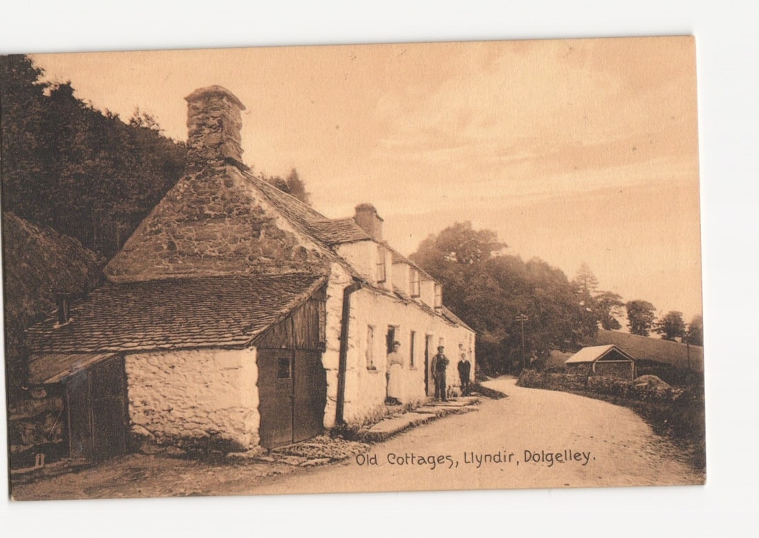 Old Cottages, Lyndir, Dolgelley, with Two Men, Early 20th Century Sepia Photographic postcard: A sepia-toned photographic image presents a collection of old stone cottages with steep slate roofs, nestled amidst dense trees under a cloudy sky. The main cottage, prominently featured, has a large 