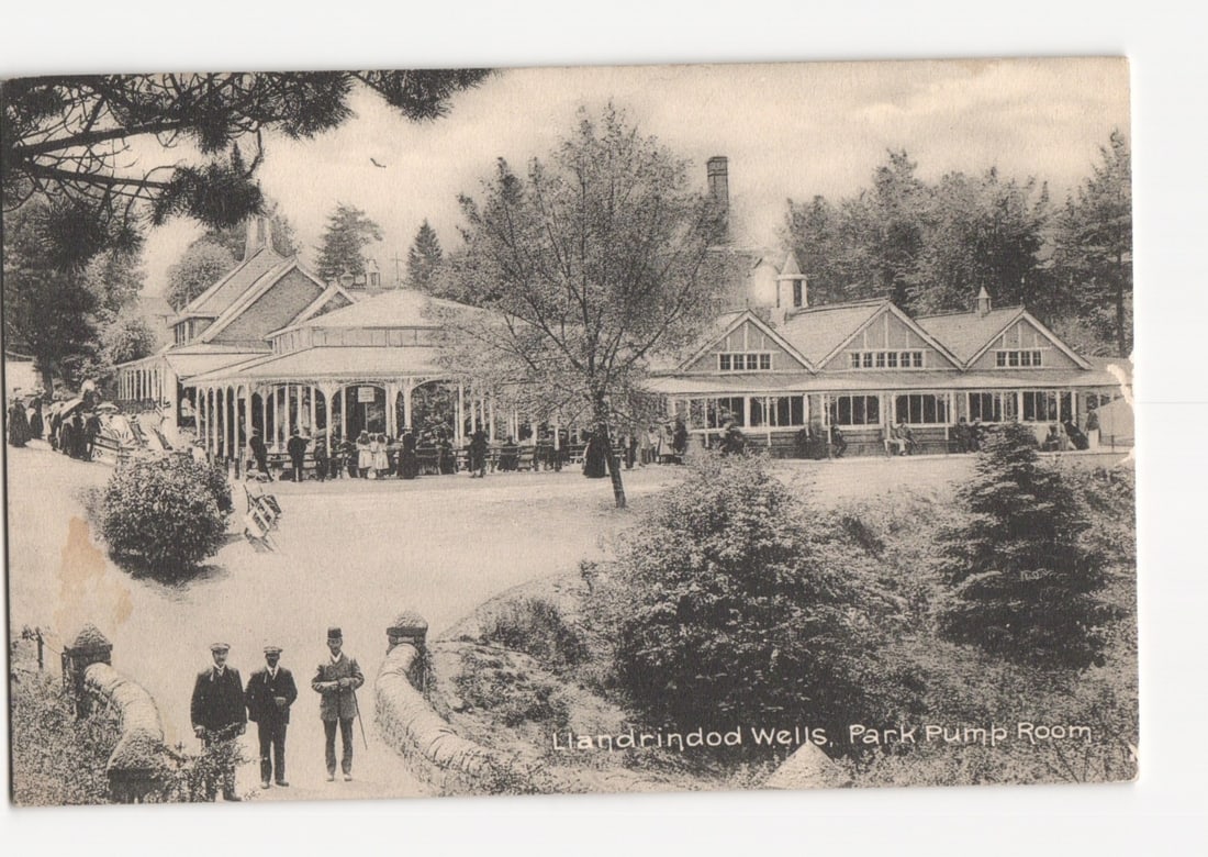 Llandrindod Wells Park Pump Room building and grounds with visitors, early 20th century postcard.: A panoramic view captures the Llandrindod Wells Park Pump Room, a multi-gabled building with an extensive veranda, situated within a landscaped park setting. To the right, another large, gabled struct