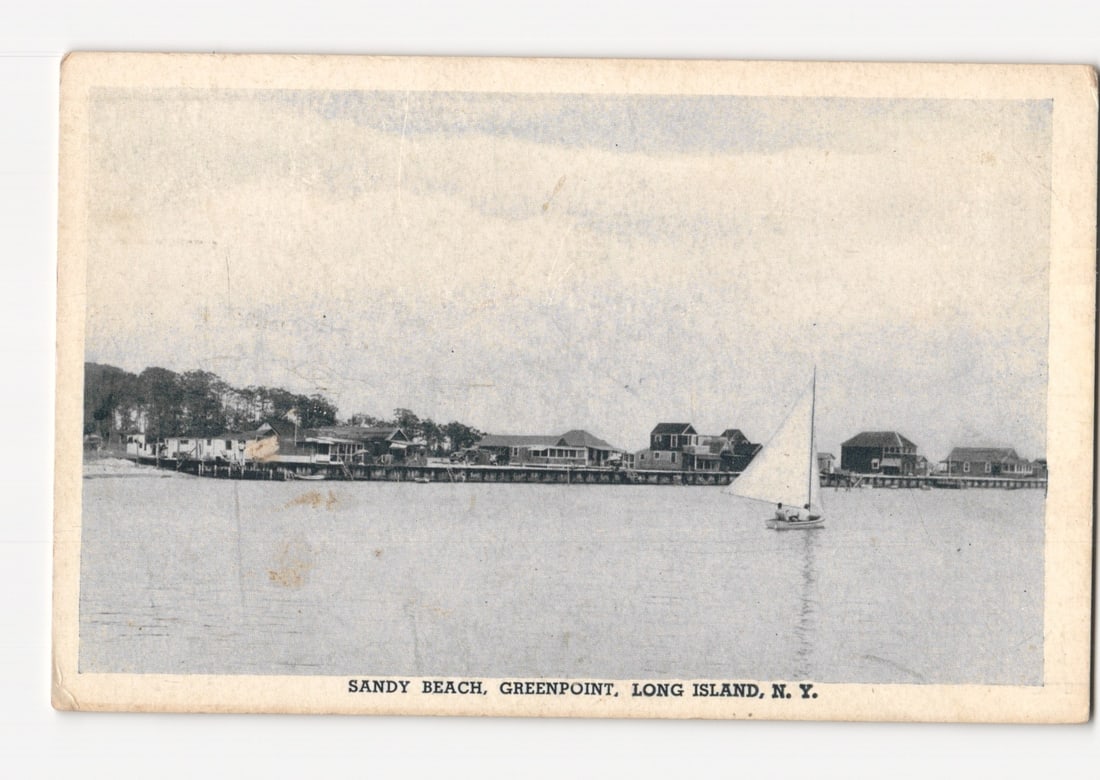 Sandy Beach, Greenpoint, Long Island, New York, with waterfront buildings and sailboat postcard: A panoramic view depicts Sandy Beach in Greenpoint, Long Island, New York. The foreground shows a calm body of water with a small sailboat, carrying two visible figures, positioned towards the center 