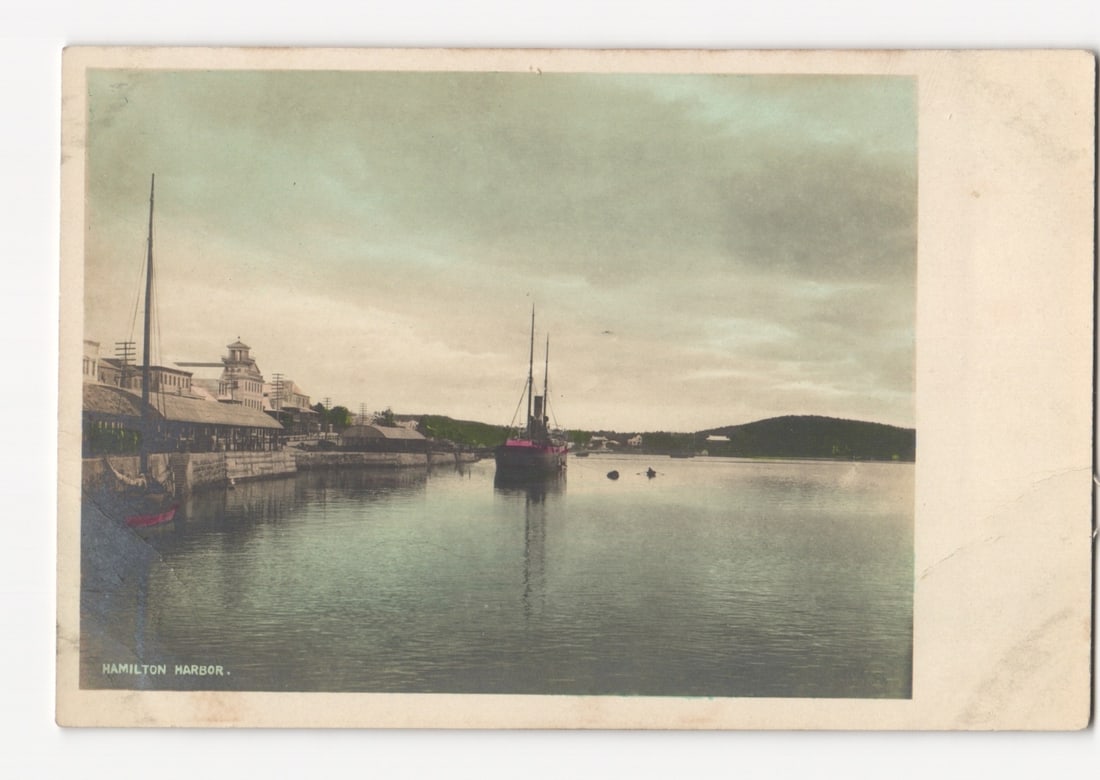 Hamilton Harbor in Bermuda with Boats and Waterfront Buildings, Early 20th Century postcard: A harbor scene features several boats on calm water, with a line of buildings along the curving shoreline under a cloudy sky. In the foreground left, a large mast from a docked vessel rises above the 