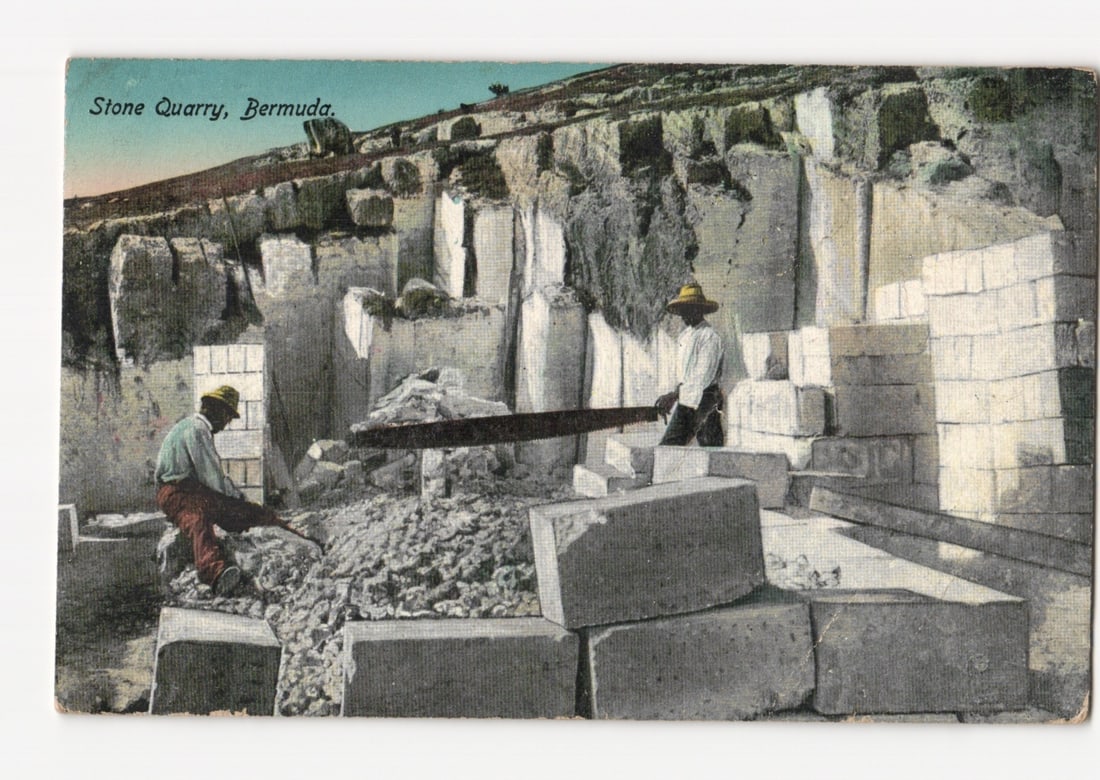 Historic postcard depicting two men laboring in the Stone Quarry, Bermuda, early 20th century.: The scene depicts two men laboring in a stone quarry, with numerous large, cut stone blocks stacked in the foreground and midground. One man, wearing a hat and light-colored shirt, kneels on the left,
