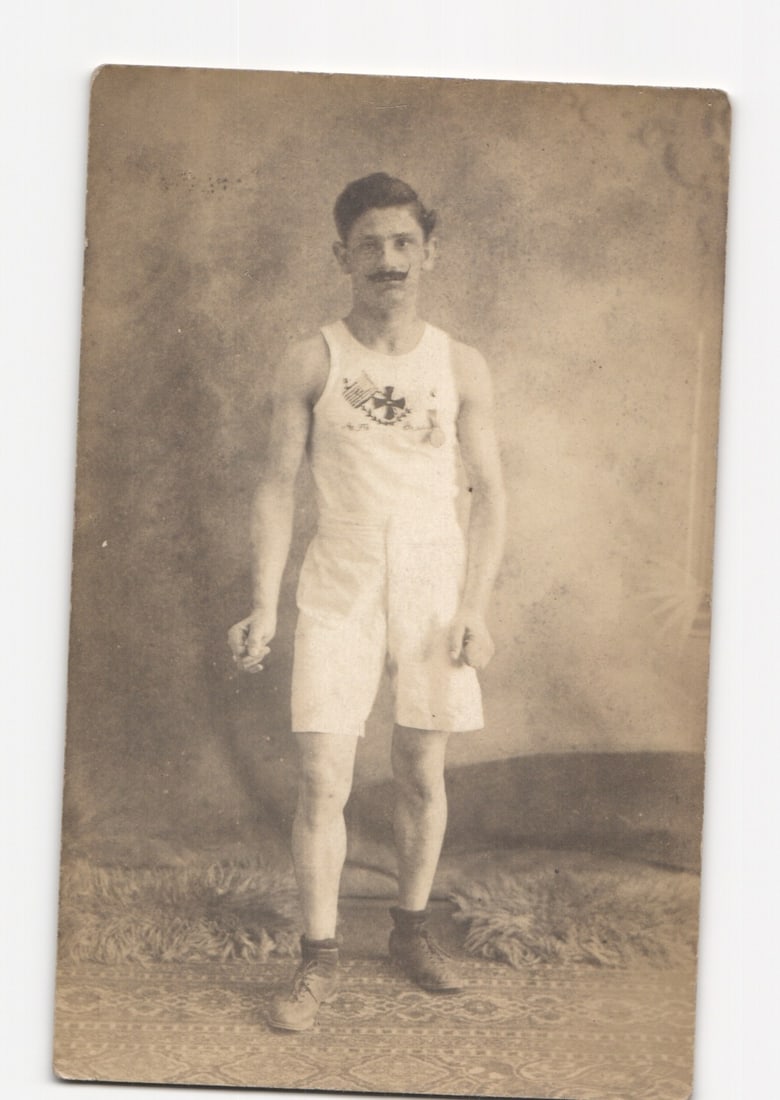 Early 20th Century Studio Portrait Postcard of a Young Man in Athletic Attire with Emblem: A full-length studio portrait photograph depicts a young man standing centrally, facing the viewer. He is dressed in a white sleeveless tank top and white shorts, with dark high-top boots. The tank to