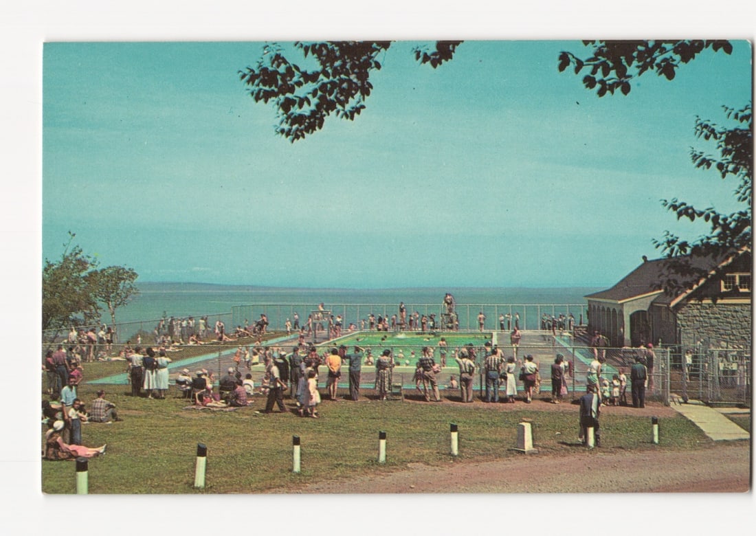 Crowded Swimming Pool and Ocean Vista, Fundy National Park, New Brunswick, Canada postcard: A color photochrome postcard depicts a large outdoor swimming pool filled with numerous bathers. The pool area, enclosed by a fence, is situated on a grassy slope overlooking a vast body of water, ext