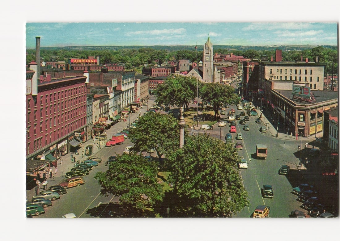 Overhead View of Public Square Looking East, Watertown, New York, Mid-20th Century postcard: An overhead view of the Public Square and a bustling street in a mid-20th century cityscape. The scene is dominated by multiple lanes of traffic and numerous vintage automobiles. Multi-story brick and
