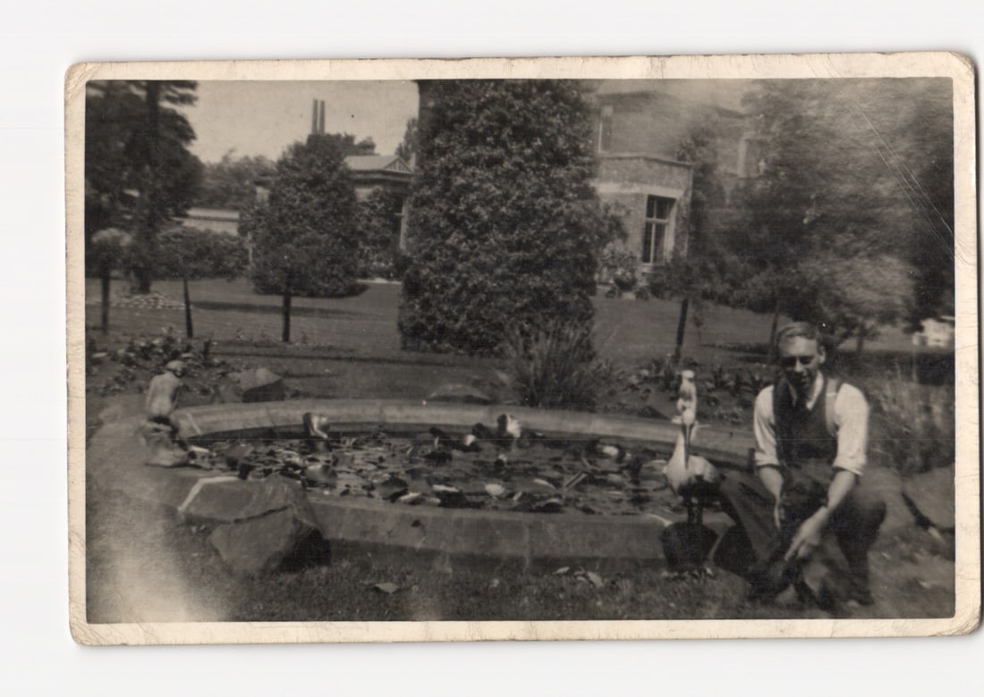 Man Posing by a Garden Pond with Ornamental Geese and Mansion in Background, Real Photo Postcard: A monochromatic photograph features a man kneeling at the lower right, positioned beside a decorative garden pond. The pond contains water lilies or similar aquatic foliage and several ornamental bird