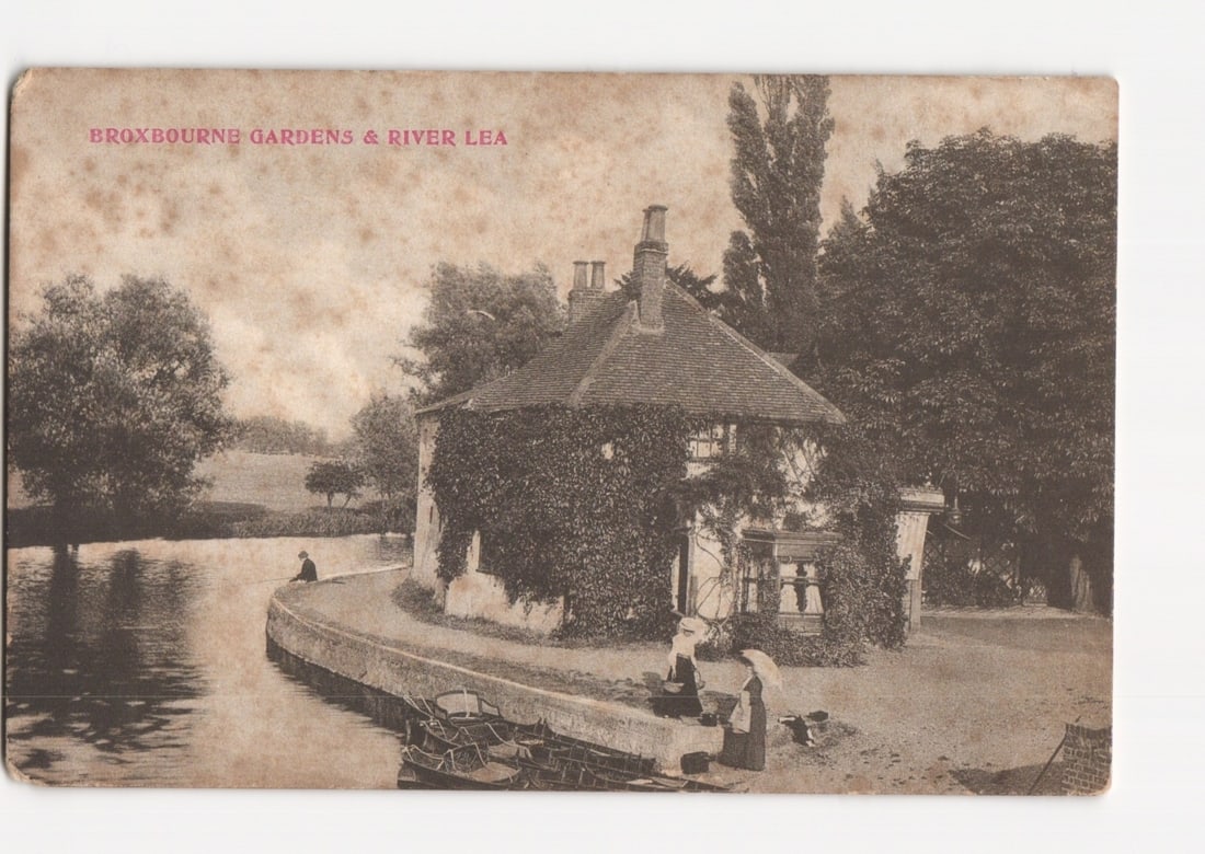 Early 20th Century postcard of Broxbourne Gardens and River Lea, featuring an ivy-covered house.: An early 20th-century halftone photographic print depicts a picturesque scene of Broxbourne Gardens and the River Lea. A charming, two-story house, heavily draped in ivy, stands prominently at the riv