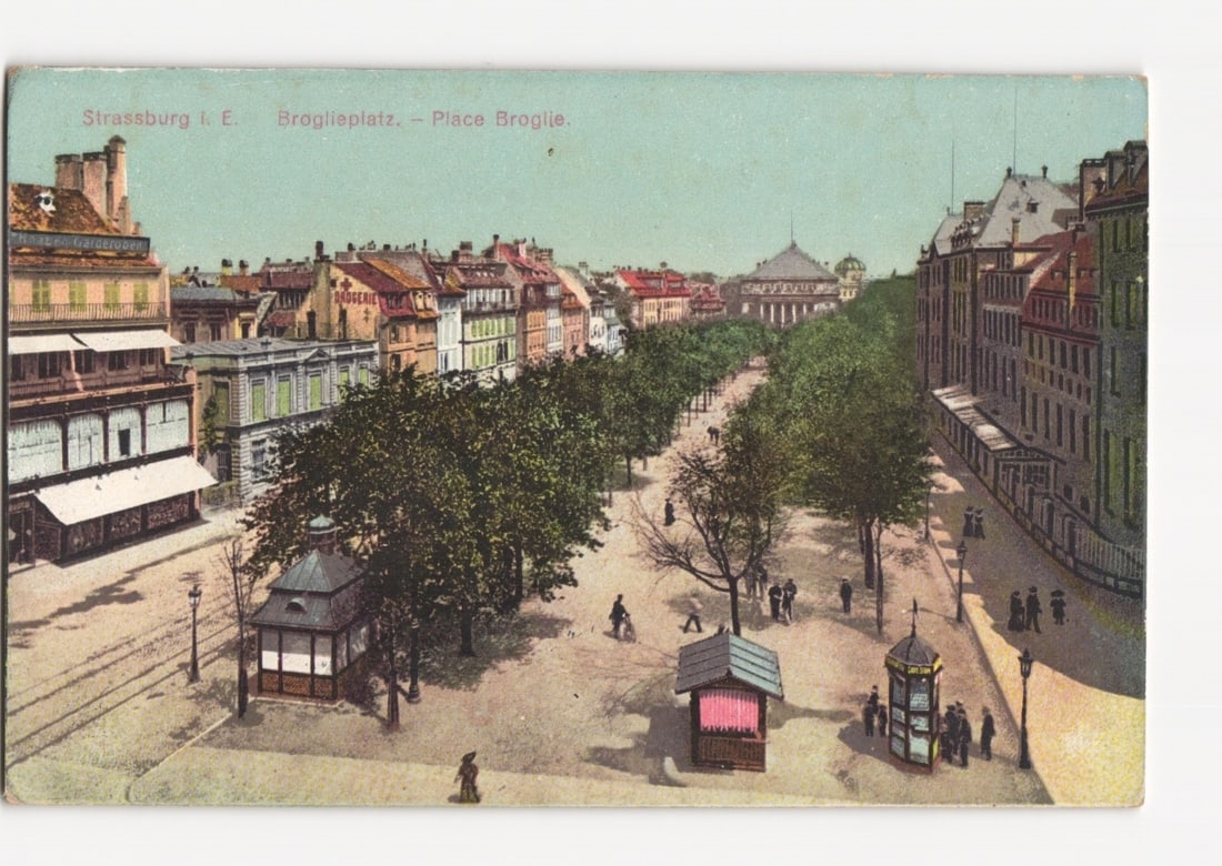 Early 20th Century Street Scene Postcard: Broglieplatz, Strassburg, with Historic Buildings: A wide panoramic view of Broglieplatz (Place Broglie) in Strassburg, featuring a tree-lined avenue receding into the background. In the foreground on the left, an open-air cafe or shop with an awning 