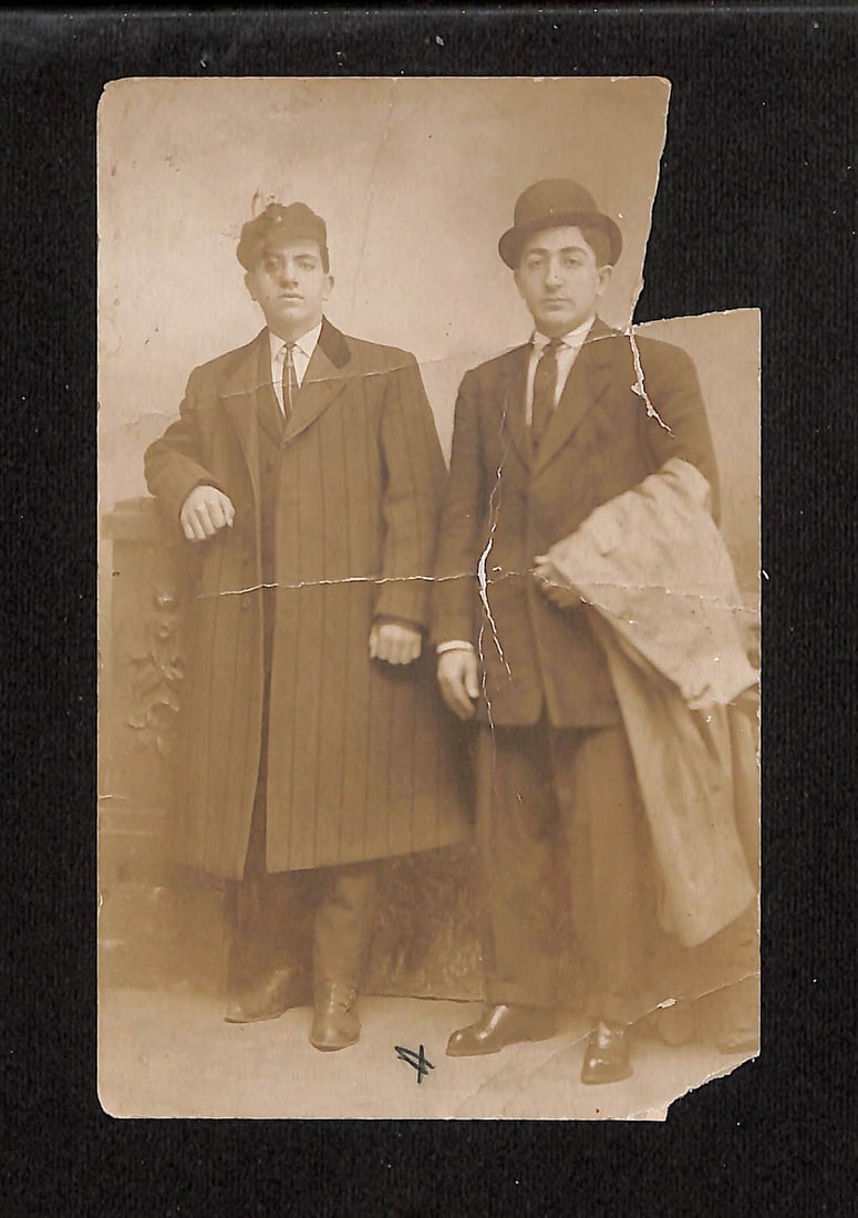 Sepia Studio Portrait of Two Young Men in Formal Attire, Early 20th Century, postcard: Full-length studio portrait of two young men. The man on the left wears a pinstripe overcoat and tie, leaning on a dark, decorative stone ledge. The man on the right wears a dark suit, tie, and bowler