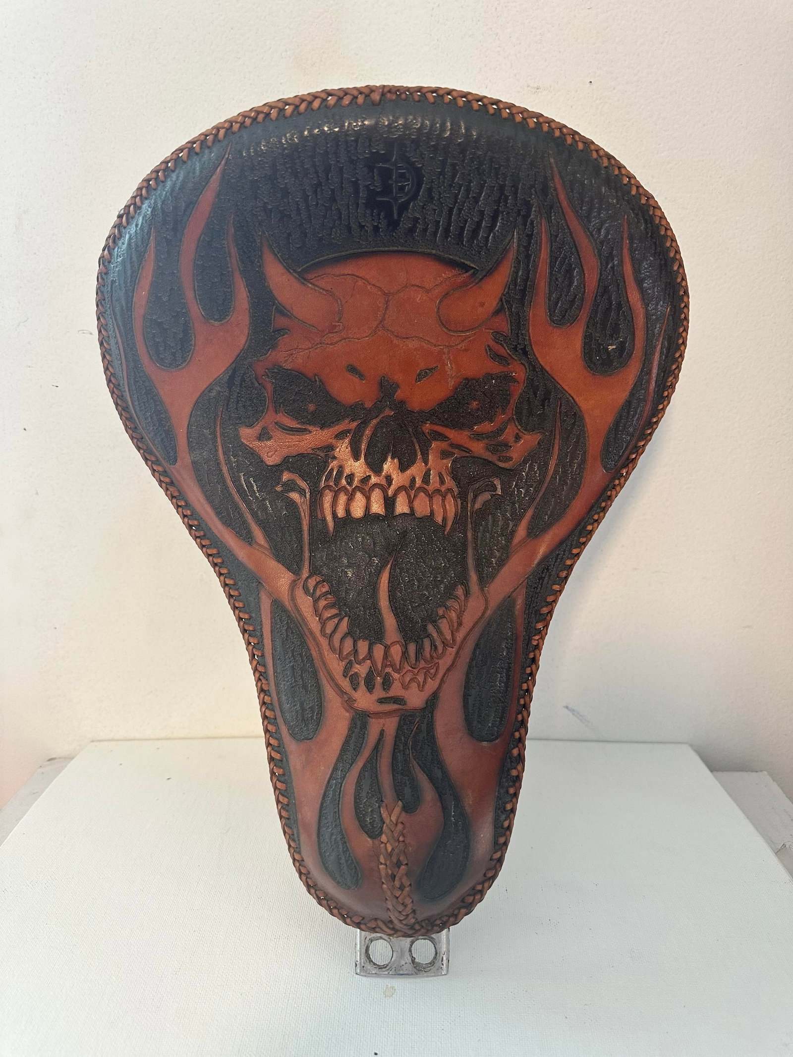 HANDMADE LEATHER SOLO SEAT: HANDMADE LEATHER SOLO SEAT FOR HARLEY. HAS LEATHER BRAIDING AROUND EDGES AND DEPICTS A FLAMING DEMON SKULL ON SEAT.