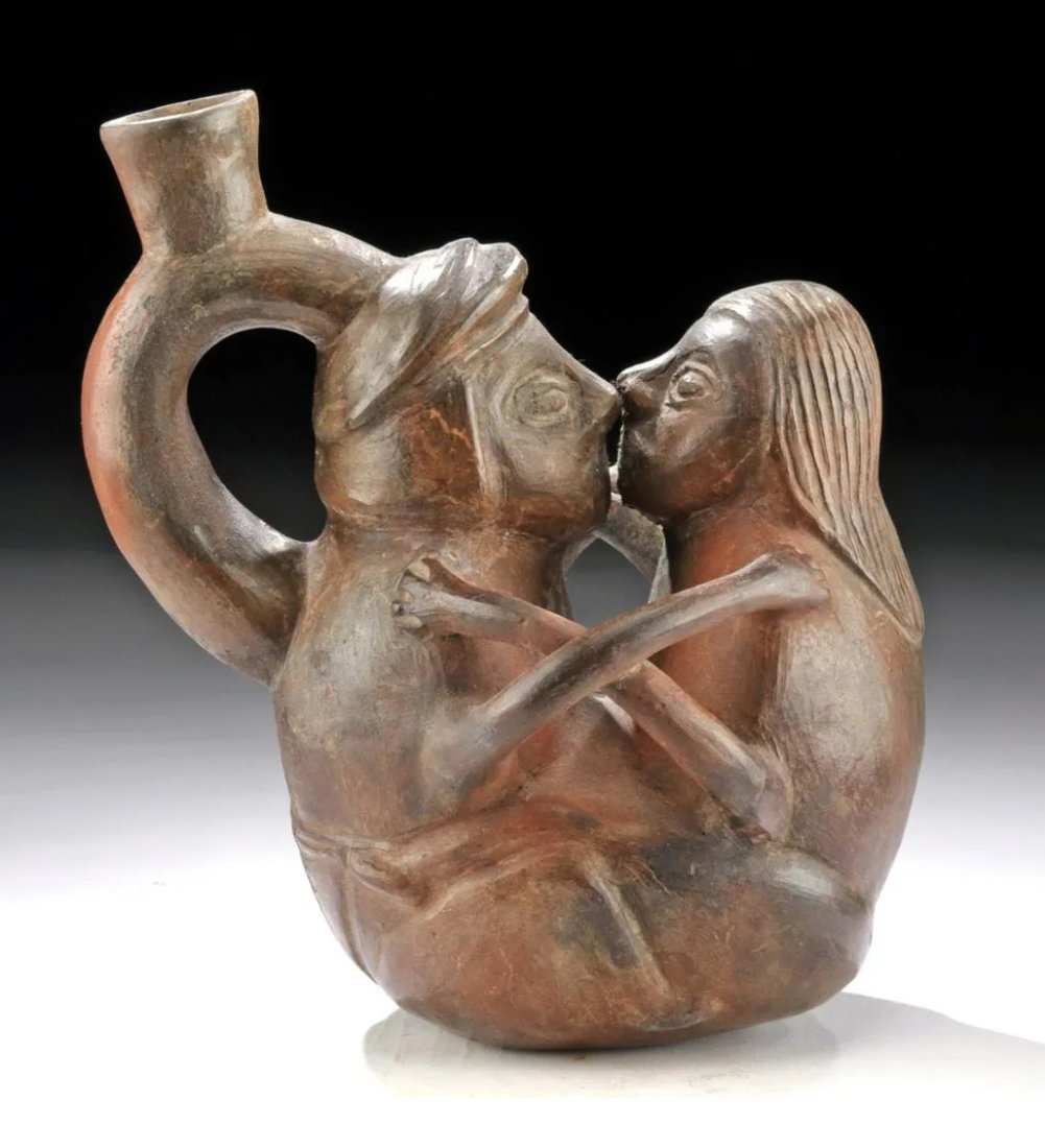Chimu Erotic Stirrup Vessel of a Couple: Ca. 900 to 1250 AD. Black ware pottery figural vessel depicting a male and female couple facing each other, kissing, and embracing; holding their hands on one another's shoulders, her raised legs rest