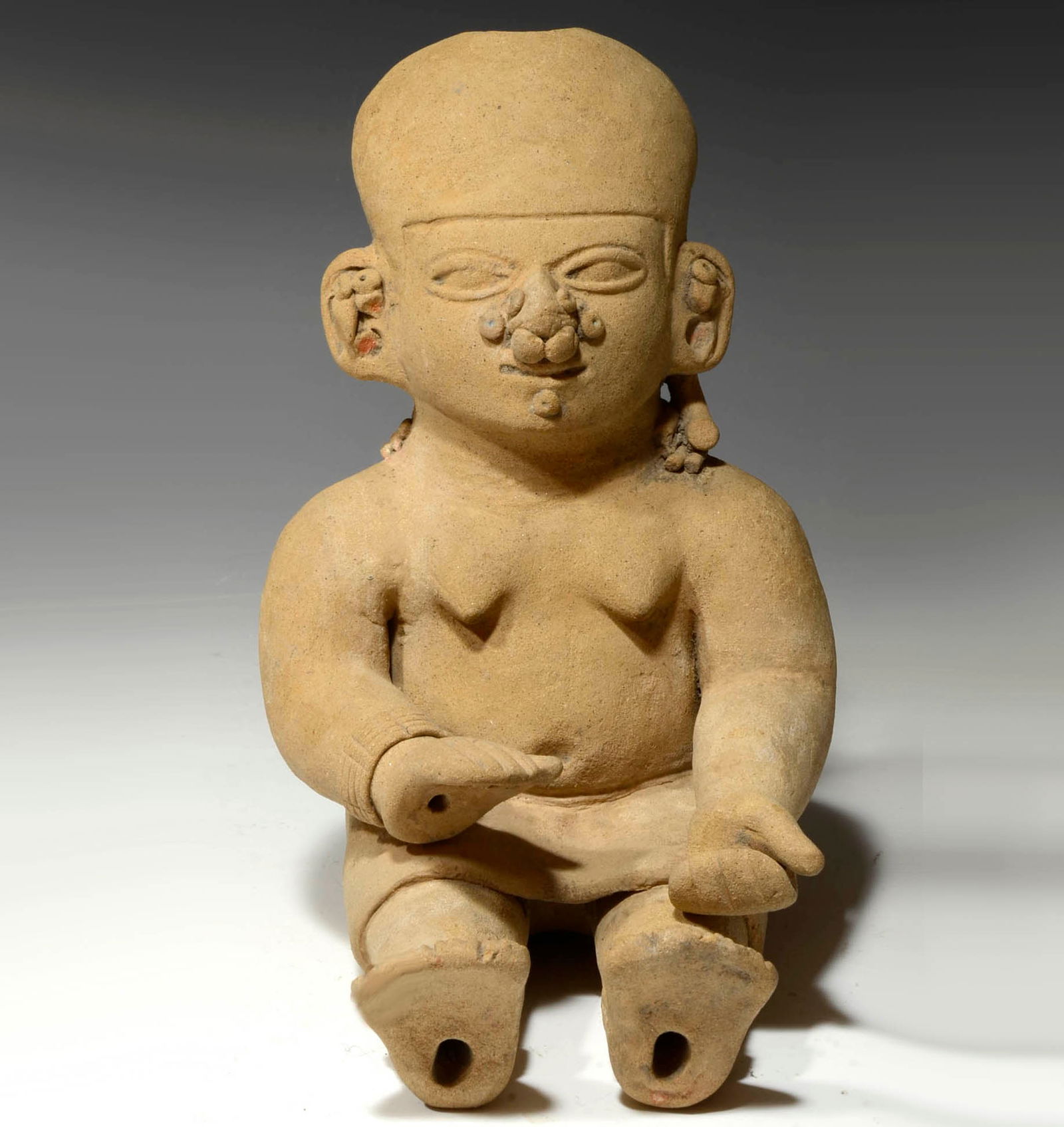 Pre-Columbian Tumaco La Tolita Seated Votive Female Figure: Ecuador, Ca 300 BC/ AD. A smooth buff color clay seated figure with arms in lap, perhaps once holding a baby, She has fine facial features and is wearing a smooth form fitted headdress and ear and nos