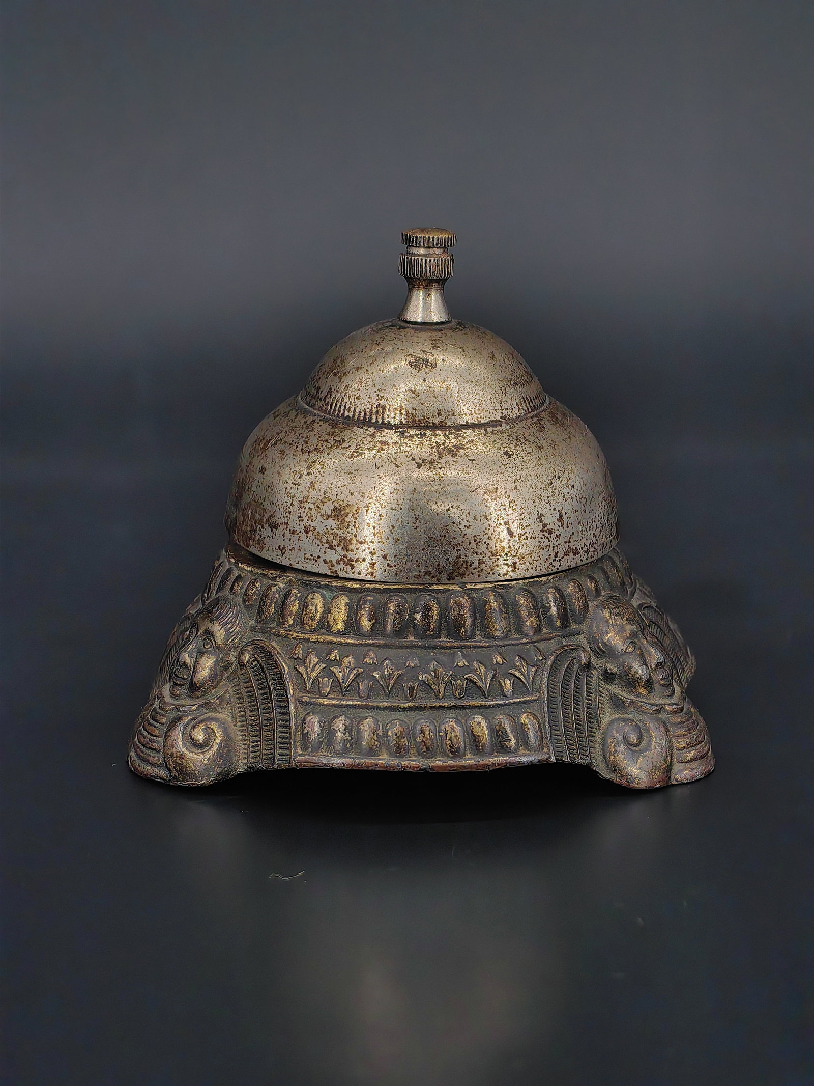 An Antique Desk Bell: A finely cast antique metal desk bell featuring a domed top with screw finial and richly decorated base. The body is adorned with classical relief motifs, including stylized winged figures and
