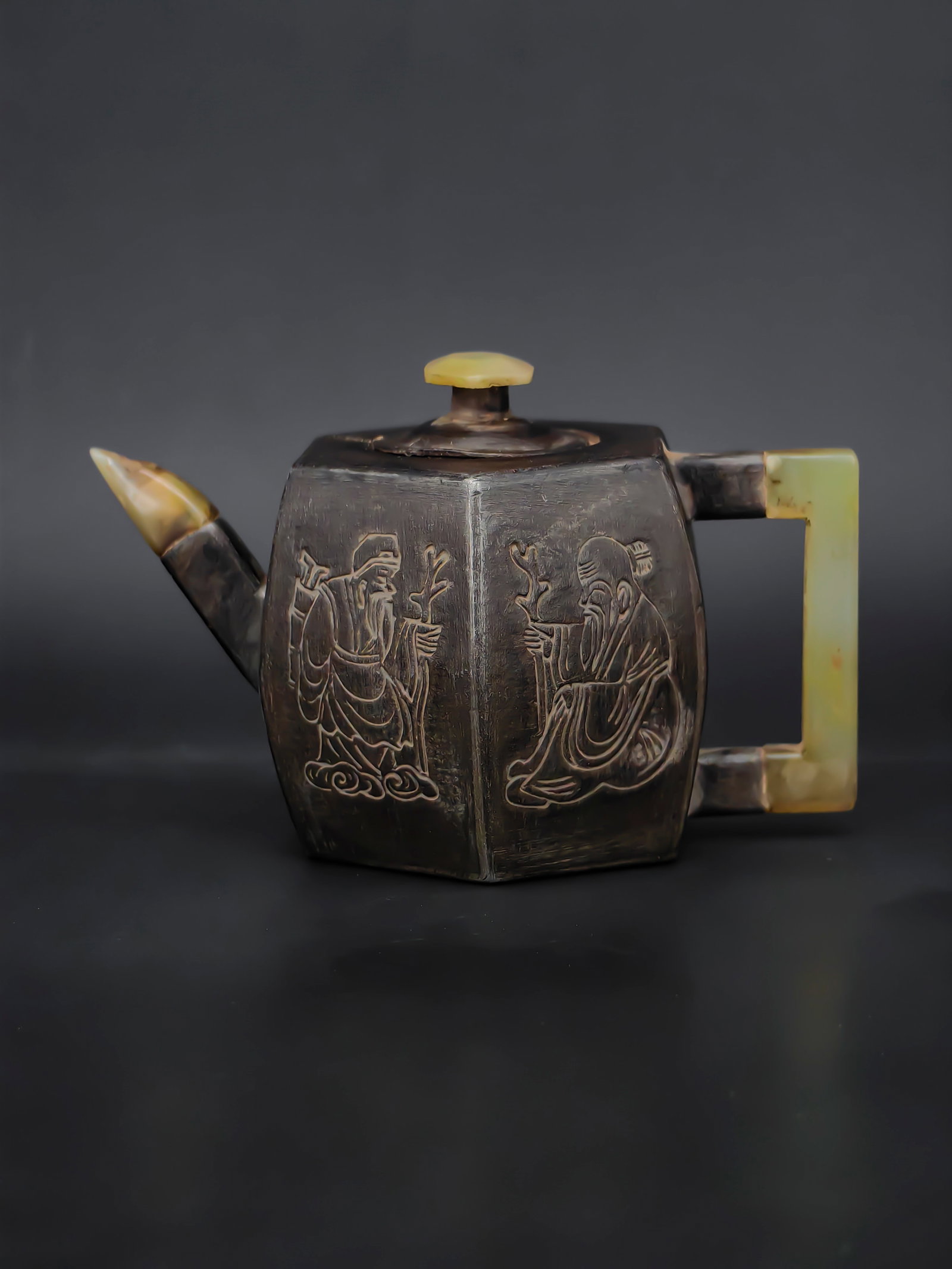 An Antique Chinese ZiSha Teapot: A finely crafted antique Yixing zisha teapot with jade-mounted handle, spout tip, and lid finial. The hexagonal body is made of dark purple clay, delicately engraved with scholar figures holding